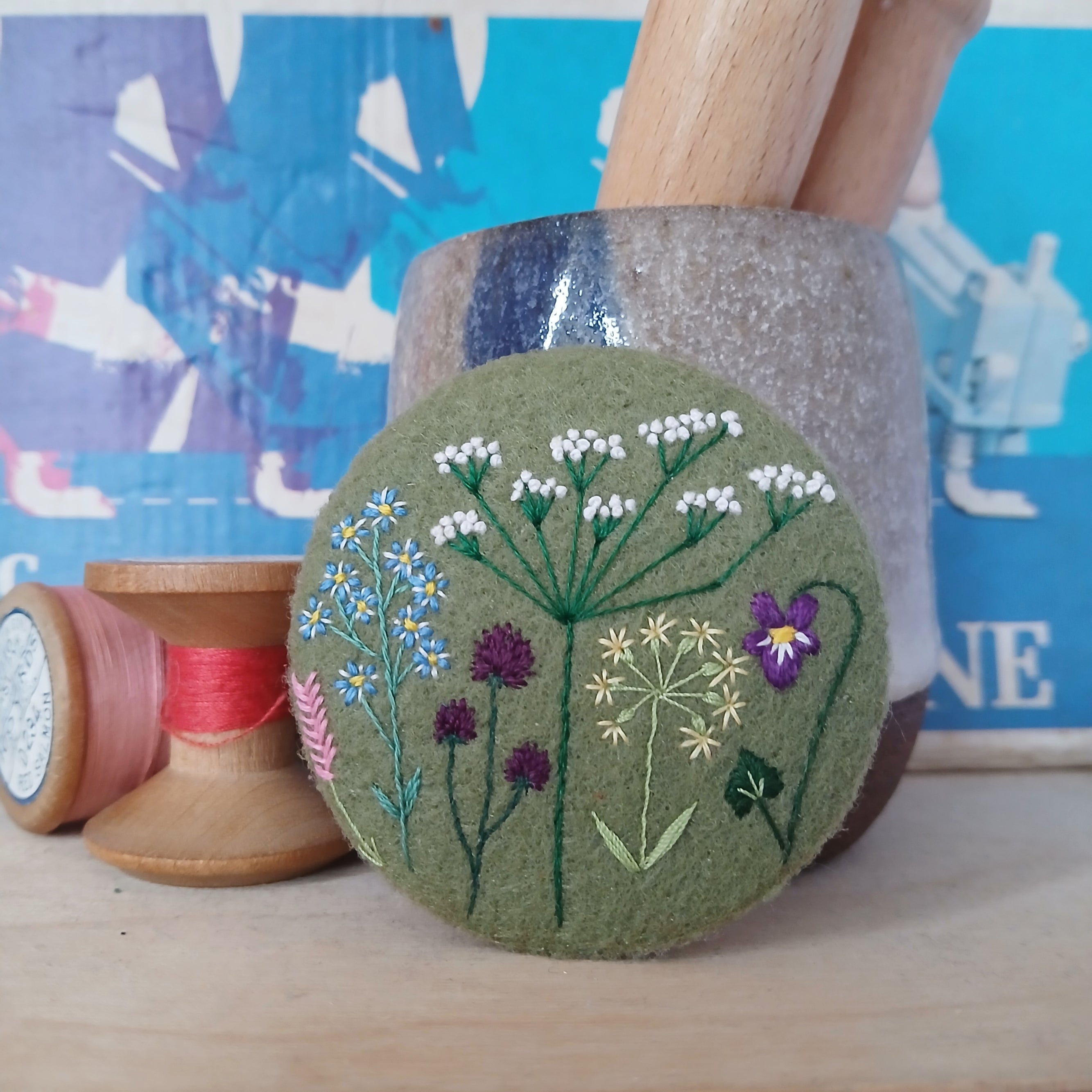A round felt pin featuring embroidered wildflowers in green, white, purple, and blue.